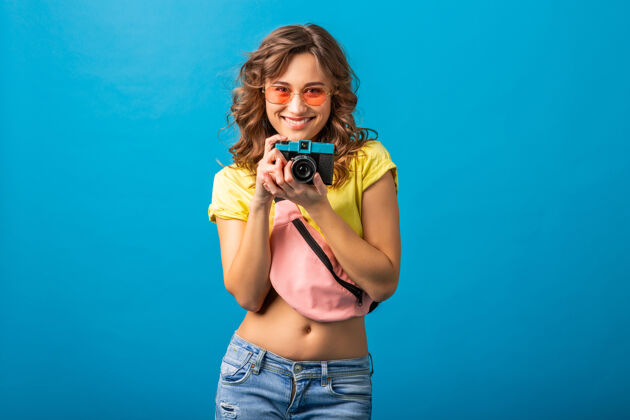 女性迷人的微笑快乐的女人摆姿势与老式相机拍照穿着时髦的夏季五颜六色服装隔离在蓝色工作室的背景年轻明亮微笑