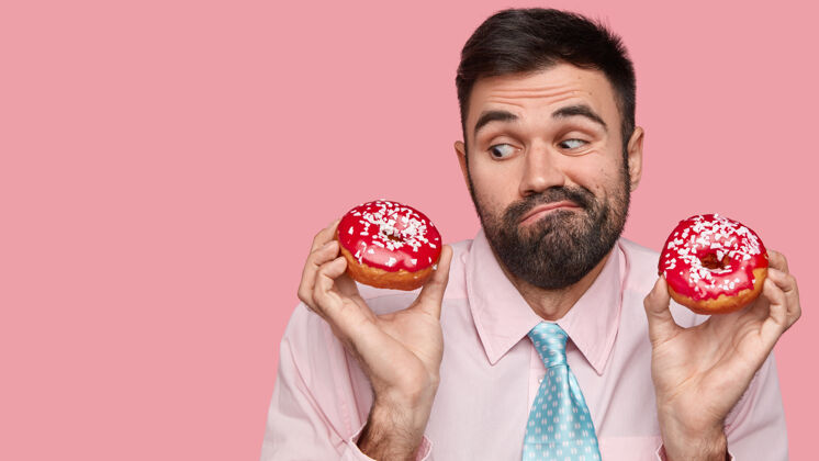 个性照片中 一个没刮胡子的白人男子穿着优雅的衣服 犹豫是否要吃垃圾食品 手里拿着甜甜圈美味领带胡茬