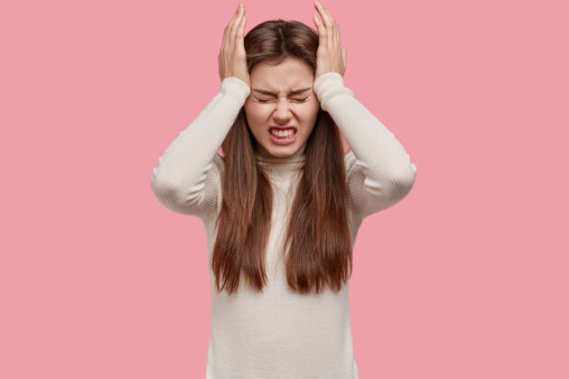 痛苦疲惫焦虑的黑发女人双手放在太阳穴上 紧咬着牙齿 头痛或偏头痛 穿着白色的衣服 隔离在粉色的背景下压力高血压偏头痛