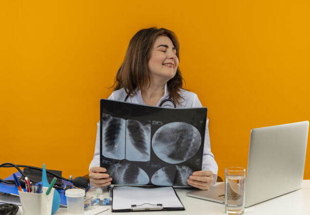 长袍闭着眼睛微笑的中年女医生穿着带听诊器的医用长袍坐在办公桌旁用笔记本电脑和医疗工具拿着x光片在隔离的橙色背景上复印笔记本电脑坐着听诊器