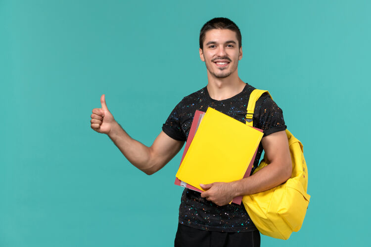 教训身穿深色t恤黄色背包的男学生正面图 浅蓝色墙上放着不同的文件快乐大学持有