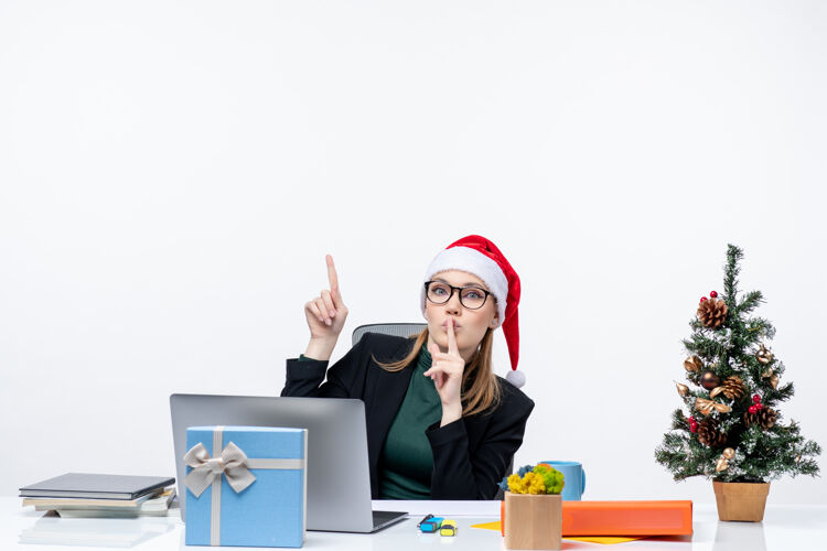 工作在白色背景的办公室里 一位好奇的商务女士戴着圣诞老人的帽子坐在一张桌子旁 桌子上放着圣诞树和礼物圣诞老人坐帽子