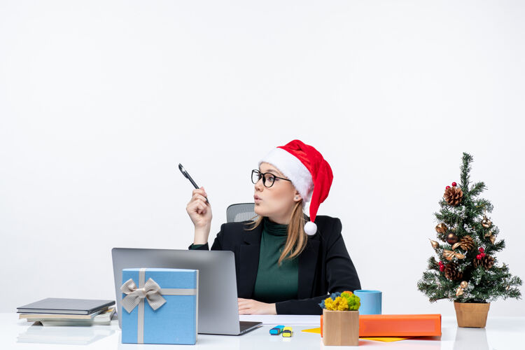 女人一个好奇的金发女人 戴着圣诞老人的帽子 坐在一张桌子旁 桌上放着一棵圣诞树和一份礼物 看着白色背景上的东西办公室金发女人什么
