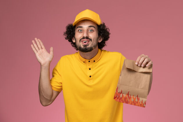 工作正面图穿着黄色制服的男快递员拿着浅粉色墙上的纸质食品包男性专业快递员