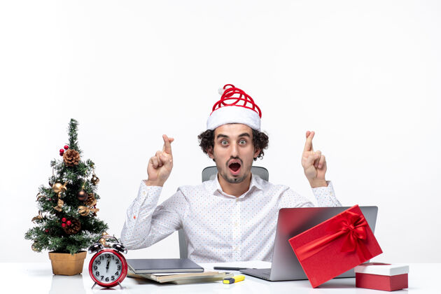 礼物快乐积极的年轻商人 戴着有趣的圣诞老人帽子 在白色背景的办公室里 手指交叉祝你好运成人人办公室
