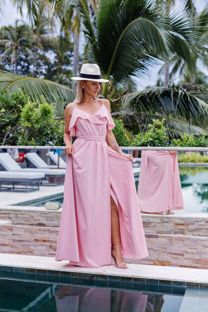 优雅穿着浪漫优雅的粉色长裙的高加索女人的Portraif在豪华别墅酒店度假 这里有令人惊叹的热带棕榈树 可以看到戴着经典白帽子的女性女性时尚配件