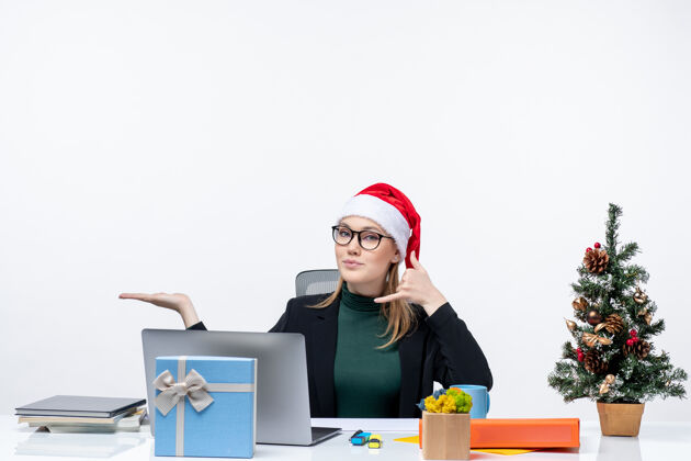 指着戴着圣诞老人帽子的金发女商人坐在一张桌子旁 桌上放着圣诞树和礼物 做着“打电话给我”的手势 在白色背景下指着办公室右边的东西商务手势商务女人
