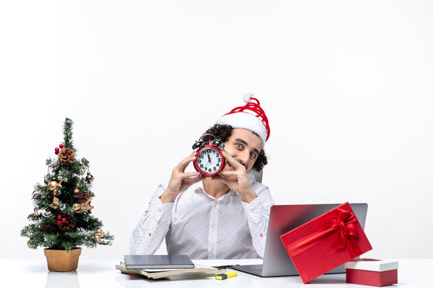 办公室年轻快乐积极的商人 戴着圣诞老人的帽子 拿着闹钟 坐在白色背景的办公室里看东西什么的年轻人坐着