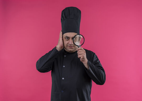 头身着厨师制服的中年男厨师手举放大镜把头放在孤立的粉红色墙上男放看