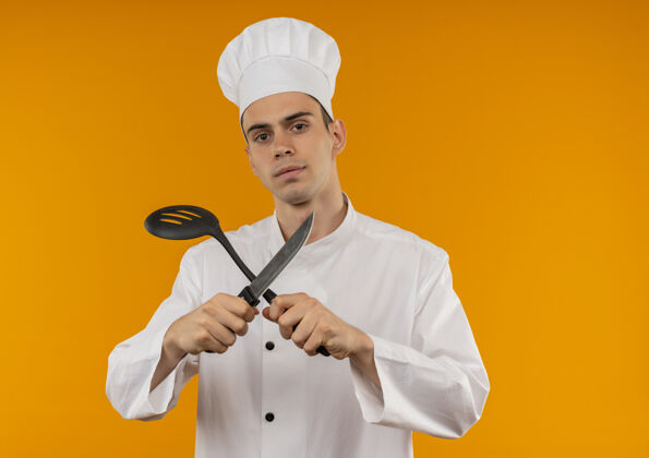 刀年轻的男酷穿着厨师制服 手里拿着勺子和刀 站在隔离的黄色墙壁上厨师看手