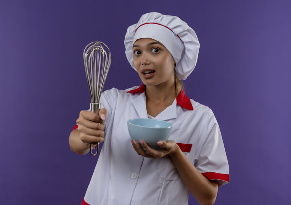 拿着年轻的女厨师穿着厨师制服 在孤零零的墙上拿着搅拌器和碗年轻厨师打