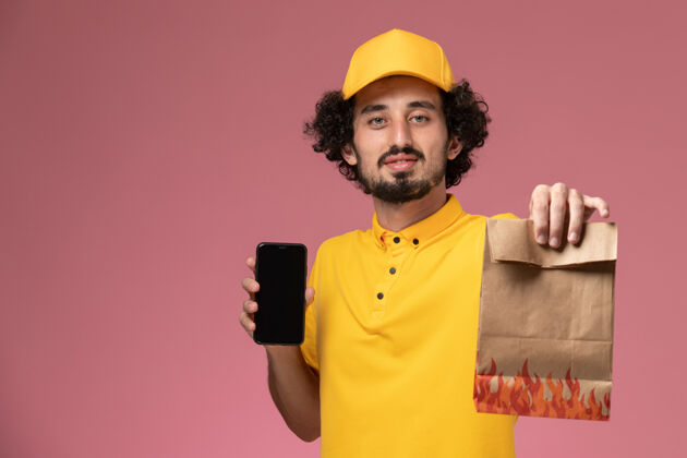 制服正面图：身穿黄色制服的男性快递员拿着食品包和智能手机站在粉红色的墙上工作男专业