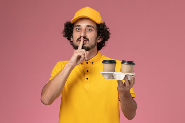 拿着正面图：身穿黄色制服 披风的男信使拿着咖啡杯在粉色墙上摆姿势男服务摆姿势