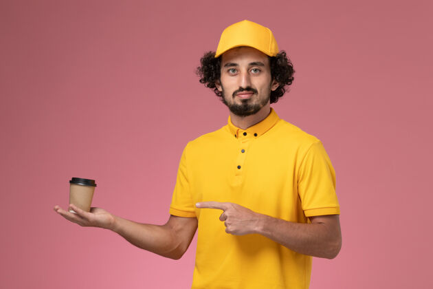 专业正面图：身穿黄色制服 披风的男性快递员在粉色墙上拿着送货咖啡杯杯子黄色工作