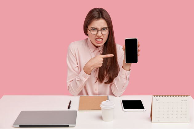 咖啡愤愤不平的年轻女子咬紧牙关 指着布洛肯手机的空屏幕 戴着圆眼镜和衬衫 拿着笔记本电脑 日历坐在工作场所恼怒工作愤怒