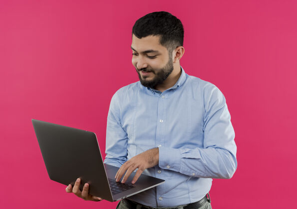 粉色身穿蓝色衬衫 留着胡须的年轻人拿着笔记本电脑 面带微笑地在上面工作笔记本电脑站着脸