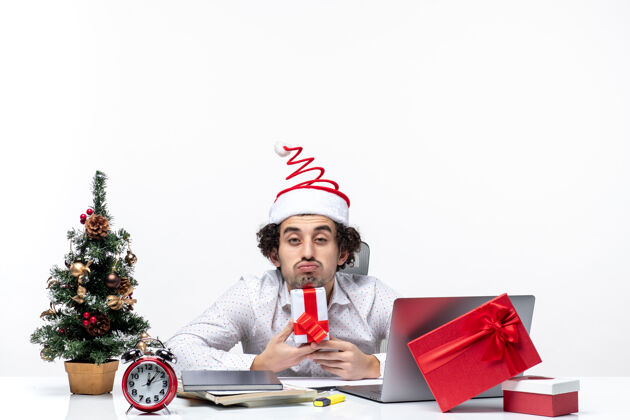 微笑圣诞节心情与忙碌的年轻商人与圣诞老人的帽子坐在办公室里 拿着他的礼物摆出白色背景的相机圣诞老人人抱着