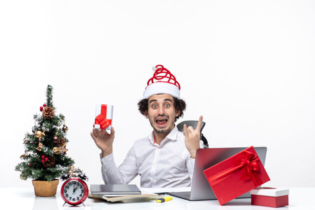 办公室新年气氛与激动情绪与年轻的商人与圣诞老人的帽子坐在办公室 并举行了他的礼物摆出白色背景的相机礼物人年轻商人