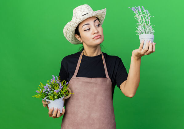 植物年轻漂亮的女园丁 围着围裙 戴着帽子 手里拿着盆栽植物 看着它们迷茫女人帽子正在