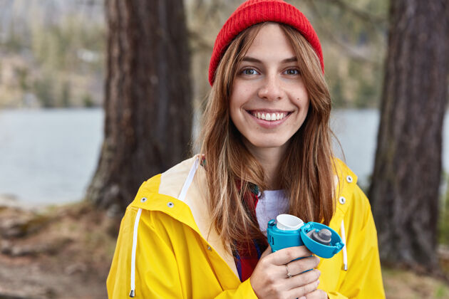 咖啡可爱的女游客在春天的森林里喝着热水瓶里的热饮 戴着红帽子 穿着黄色雨衣 对着镜头灿烂地笑着拍摄野餐野营