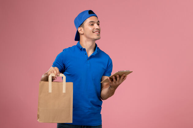 专业正面图：身穿蓝色制服和斗篷的年轻男性快递员 手上拿着快递包裹和记事本放在粉红色的墙上人制服手