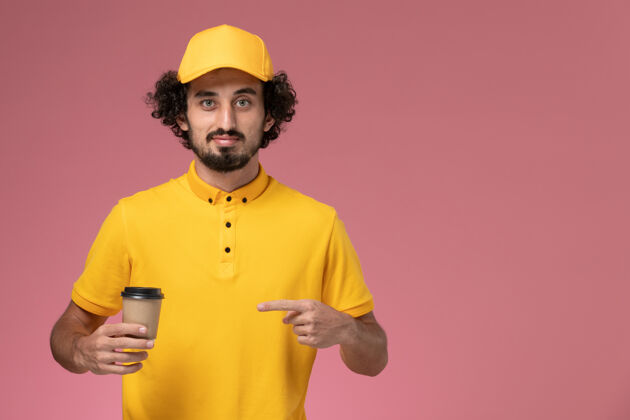 咖啡正面图：身穿黄色制服 披风的男性快递员在粉色墙上拿着送货咖啡杯送货视野职业
