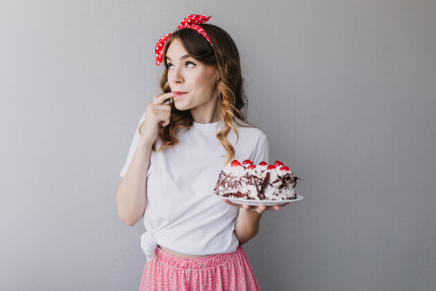 快乐可爱的卷发女孩品尝草莓蛋糕室内拍摄浪漫的女模特 头发上系着红丝带 手里拿着美味的馅饼生日休闲人
