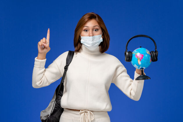风景正面图身穿白色球衣的年轻女学生戴着面具 背着背包 拿着地球仪 蓝色墙上挂着耳机背包人地球环球