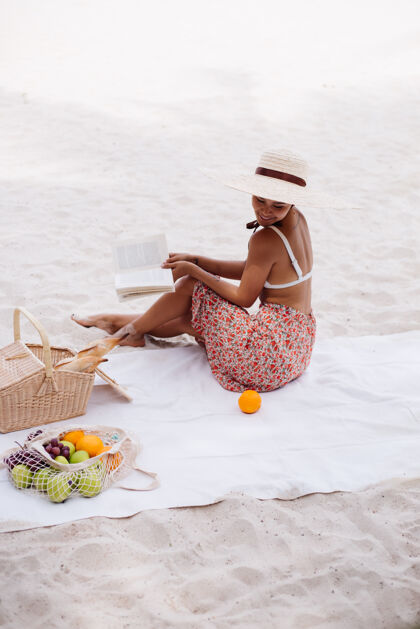 户外一个年轻女子戴着草帽 穿着白色针织衣服坐在沙滩地毯上休闲奢华野餐