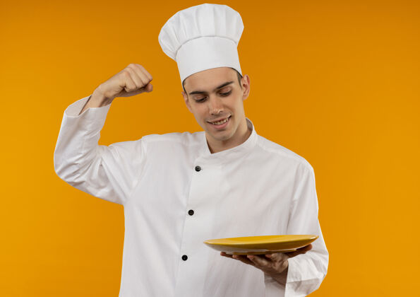 高兴高兴的年轻男酷穿着厨师制服看着盘子显示出强烈的姿态手势盘子展示