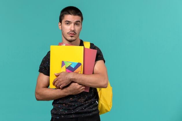持有前视图中的男学生穿着深色t恤黄色背包 在浅蓝色的墙上拿着文件和抄写本快乐背包学院