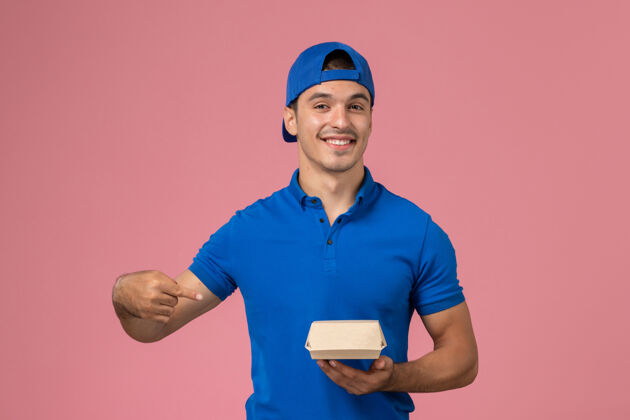 人正面图：身穿蓝色制服披肩的年轻男性快递员手持粉色墙上的快递食品包员工送货快餐
