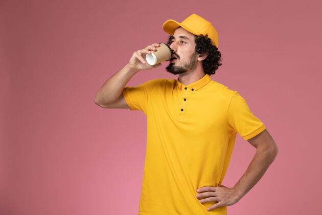 肖像正面图穿着黄色制服的男信使和披风在粉红色的墙上喝咖啡漂亮咖啡送货
