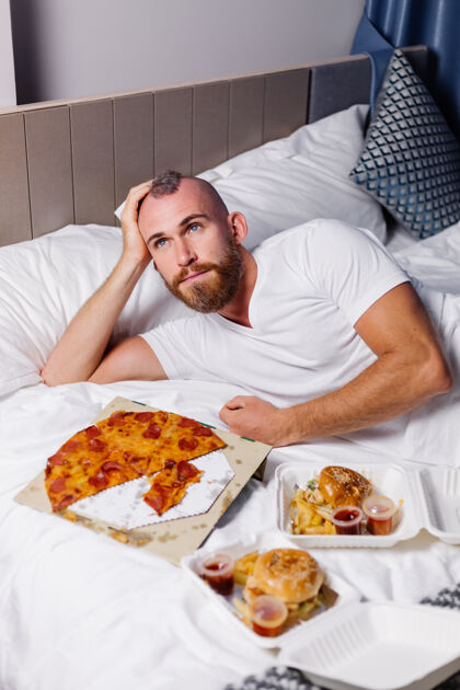 男人快乐的白种人在家里卧室里吃快餐在床上男人在网上叫外卖吃比萨饼和汉堡在舒适的房间卧室咬帅哥