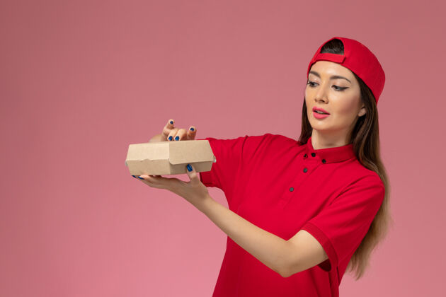 漂亮正面图：身穿红色制服和斗篷的年轻女快递员 手上拿着一个小小的快递食品包 放在粉红色的墙上快餐制服红色