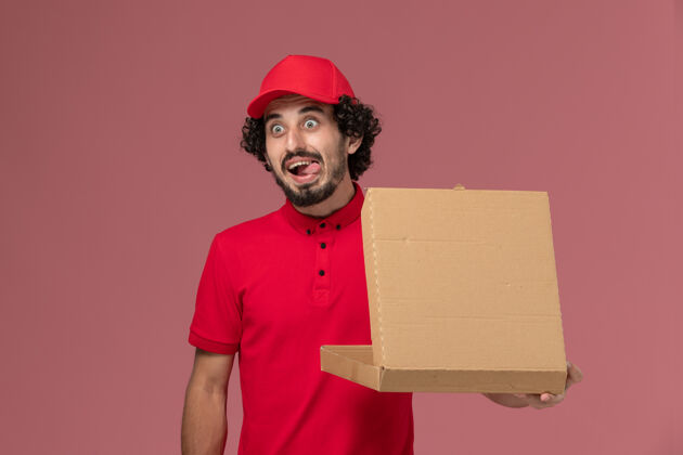 视图正面图：身穿红色衬衫和披风的男性快递员手持浅粉色墙上的送货食品箱人帽子送货