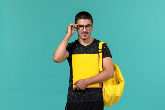 男学生身穿深色t恤黄色背包的男学生正面图 浅蓝色墙上放着不同的文件持有档案人