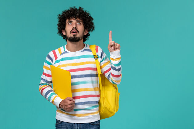 正面身穿条纹衬衫 背着黄色背包 在蓝色墙上拿着文件的男学生的正视图教训大学男