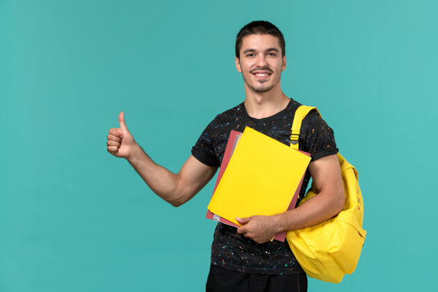 教训身穿深色t恤黄色背包的男学生正面图 浅蓝色墙上放着不同的文件快乐大学持有