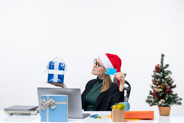 电脑积极的女商人戴着圣诞老人的帽子 戴着眼镜坐在一张桌子旁 手里拿着圣诞礼物和银行卡在办公室股票的形象礼物股票办公室