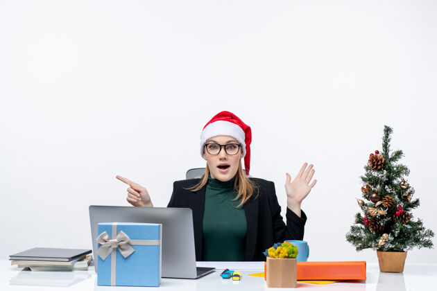 震惊一位戴着圣诞老人帽子的商务女士坐在一张桌子旁 桌子上放着圣诞树和礼物 她非常震惊 她仔细地注视着白色背景上的一些东西工作东西商务女士