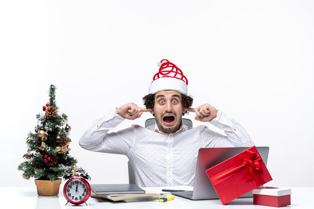 圣诞老人愤怒的年轻商人戴着滑稽的圣诞老人帽子庆祝圣诞节 他闭上耳朵 在白色背景的办公室里阻止别人聆听防止办公室听