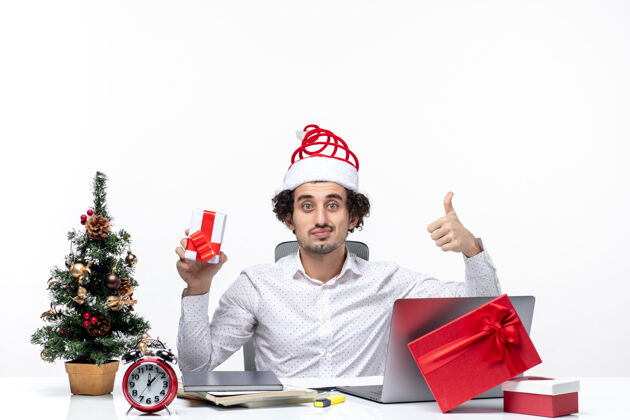 男性圣诞气氛与微笑自豪的年轻商人圣诞老人帽子和举行他的礼物指着上面的白色背景年轻指着商人