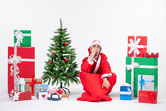 困惑困惑兴奋的年轻人打扮成圣诞老人 拿着礼物和装饰好的圣诞树坐在白色背景的地上礼物圣诞树装饰