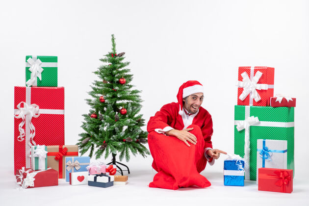 人情绪激动的年轻人打扮成圣诞老人 拿着礼物和装饰好的圣诞树坐在白色背景的地上圣诞树圣诞老人圣诞老人