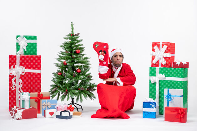 悲伤悲伤的圣诞老人坐在地上 穿着圣诞袜 靠近礼物和装饰在白色背景上的新年树坐装饰袜子