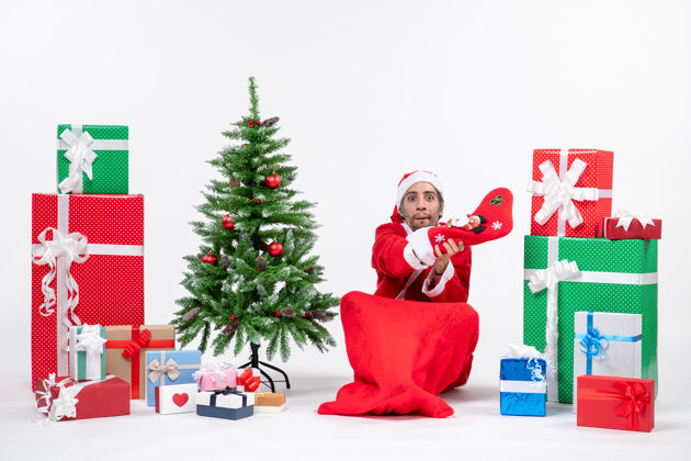 圣诞老人新年心情惊喜的圣诞老人坐在地上 穿着圣诞袜 在礼物旁边装饰圣诞树 背景是白色的礼物圣诞老人圣诞