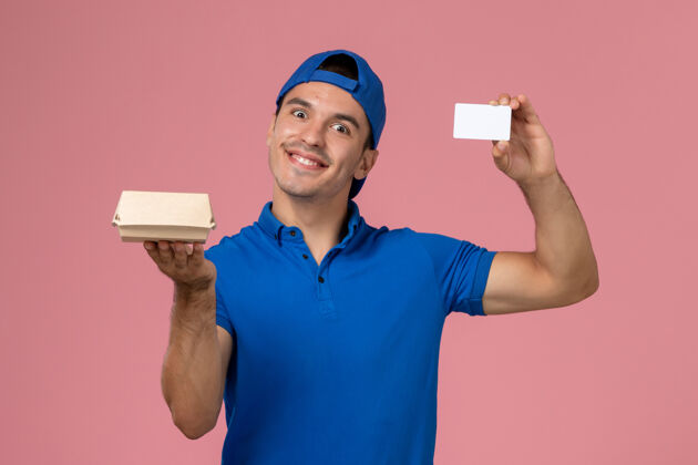 年轻男快递正面图：身穿蓝色制服披肩的年轻男性信使手拿着小快递食品包和浅粉色墙上的白色卡片年轻人男粉色