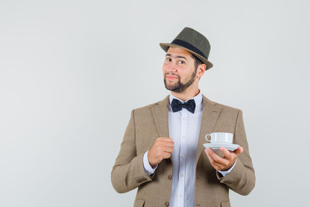 男人年轻人穿着西装 戴着帽子 手里拿着白色的茶杯和茶碟 看上去很开朗 正对着窗外成人年轻看