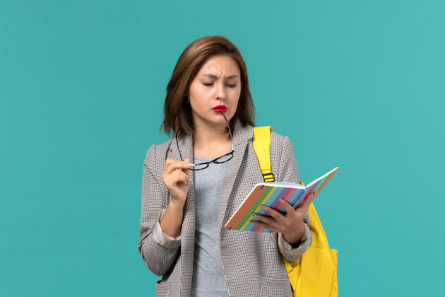 学校身穿灰色夹克 背着黄色书包 拿着抄写本 在浅蓝色墙上看书的女学生的正视图夹克穿戴阅读